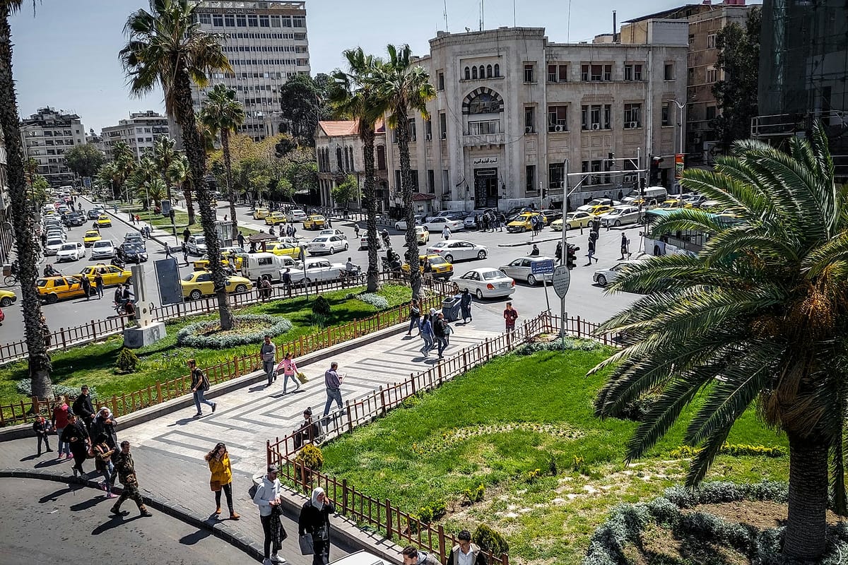 ساحة في وسط دمشق فيها نخيل وسيارات وناس يعبرون.